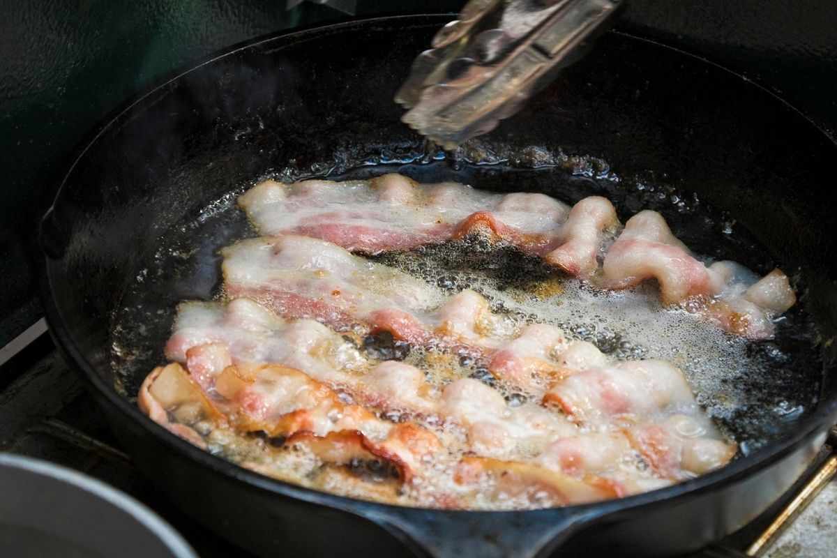 Five strips of bacon cooking in a large iron skillet.