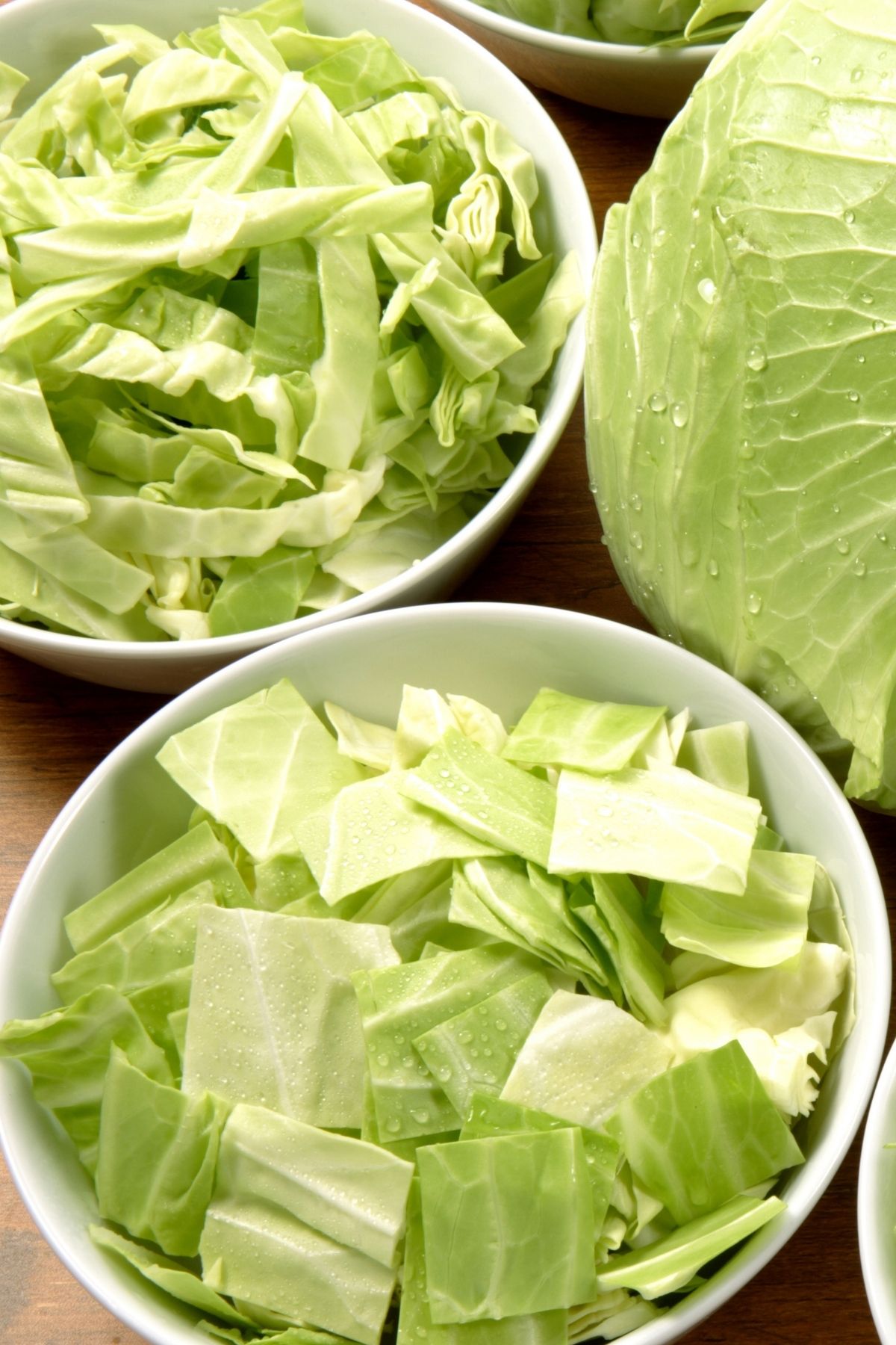 Roughly chopped cabbage placed into two different bowls with a whole head of cabbage in the background.