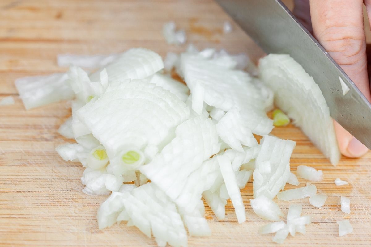 A small onion being finely chopped with a knife on a wooden cutting board.