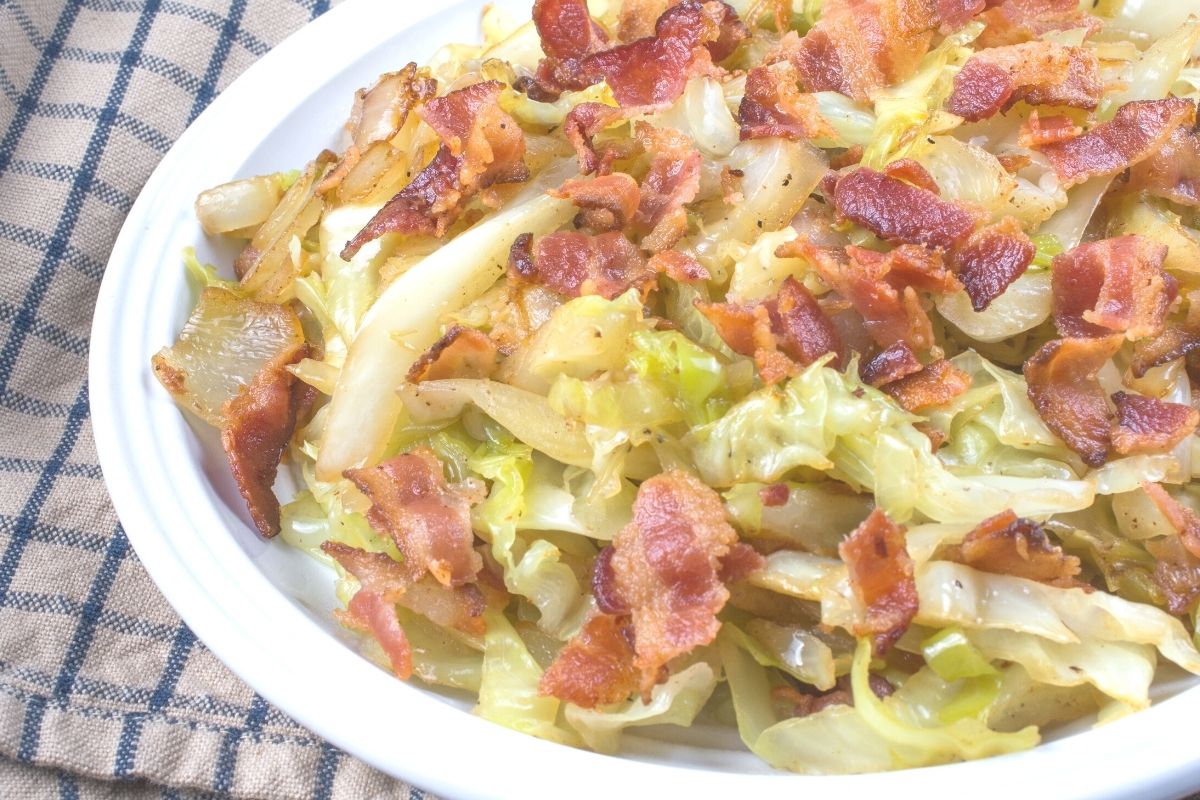 A bowl of southern fried cabbage with bacon in a white bowl resting on a kitchen towel.