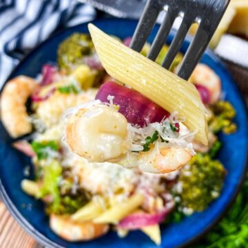 a plate of garlic shrimp pasta with a single bite of shrimp, penne, and red onion on a fork in the foreground.