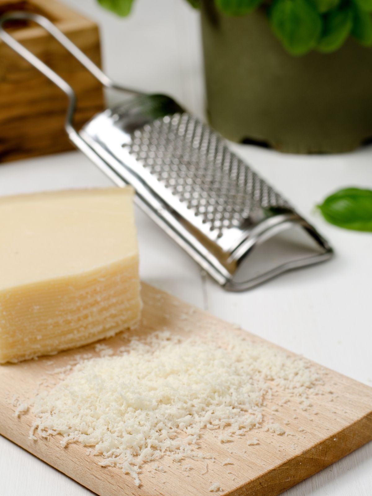 A block of parmesan cheese resting on a cutting board. Some of the cheese has been shredded in the foreground and a hand-held cheese grater is in the background.