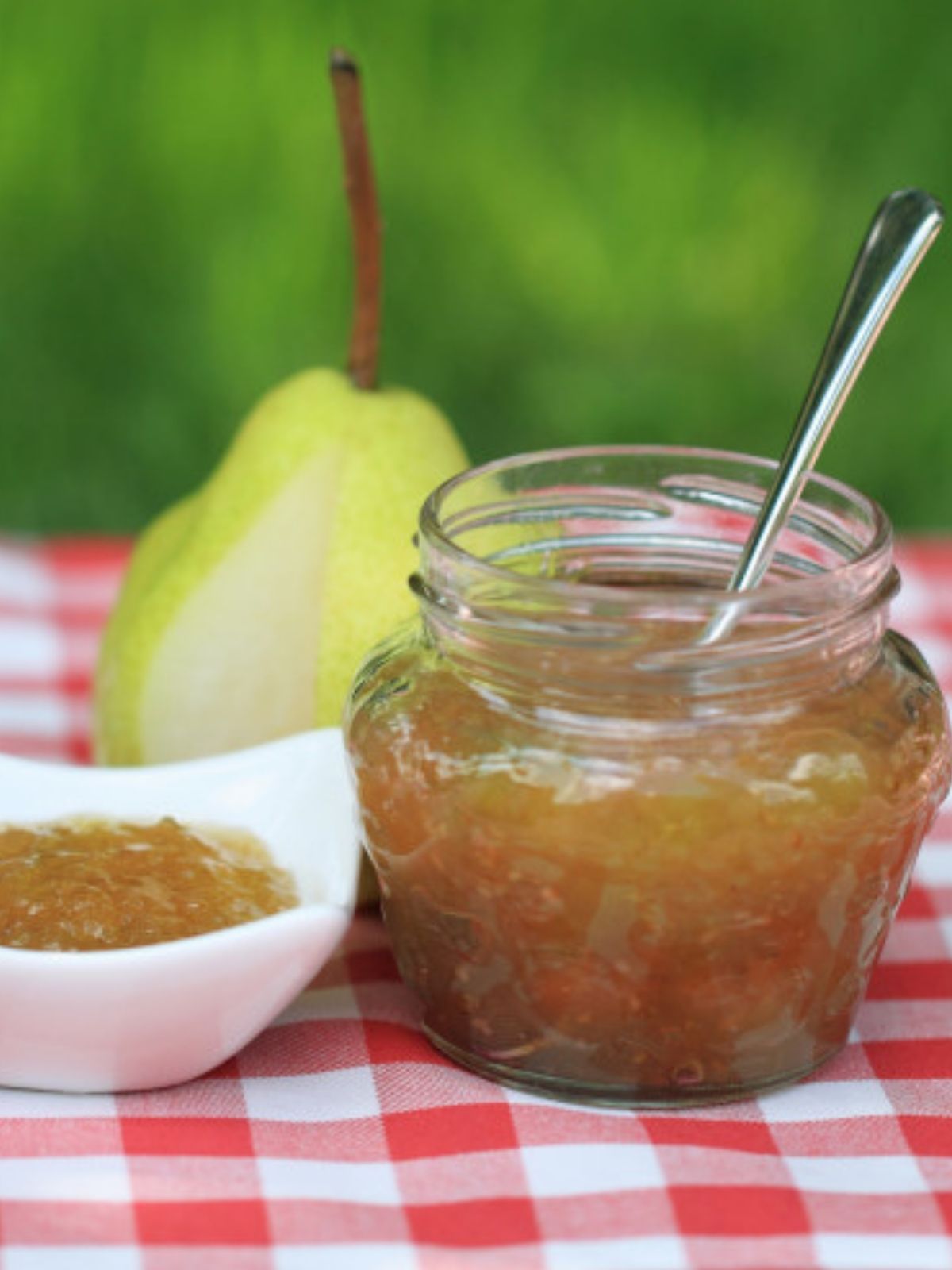 an open jar of pear butter with a silver spoon dipped inside. A fresh pear rests in  the background.