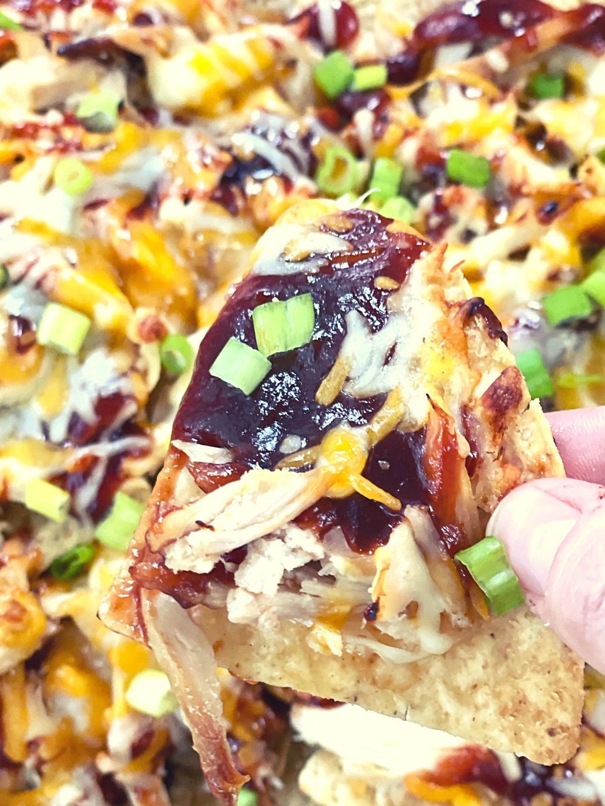 a sheet pan of baked nachos in the background with a single chip held in the foreground. The nacho has BBQ sauce, cheese, chicken, and green onion on it.