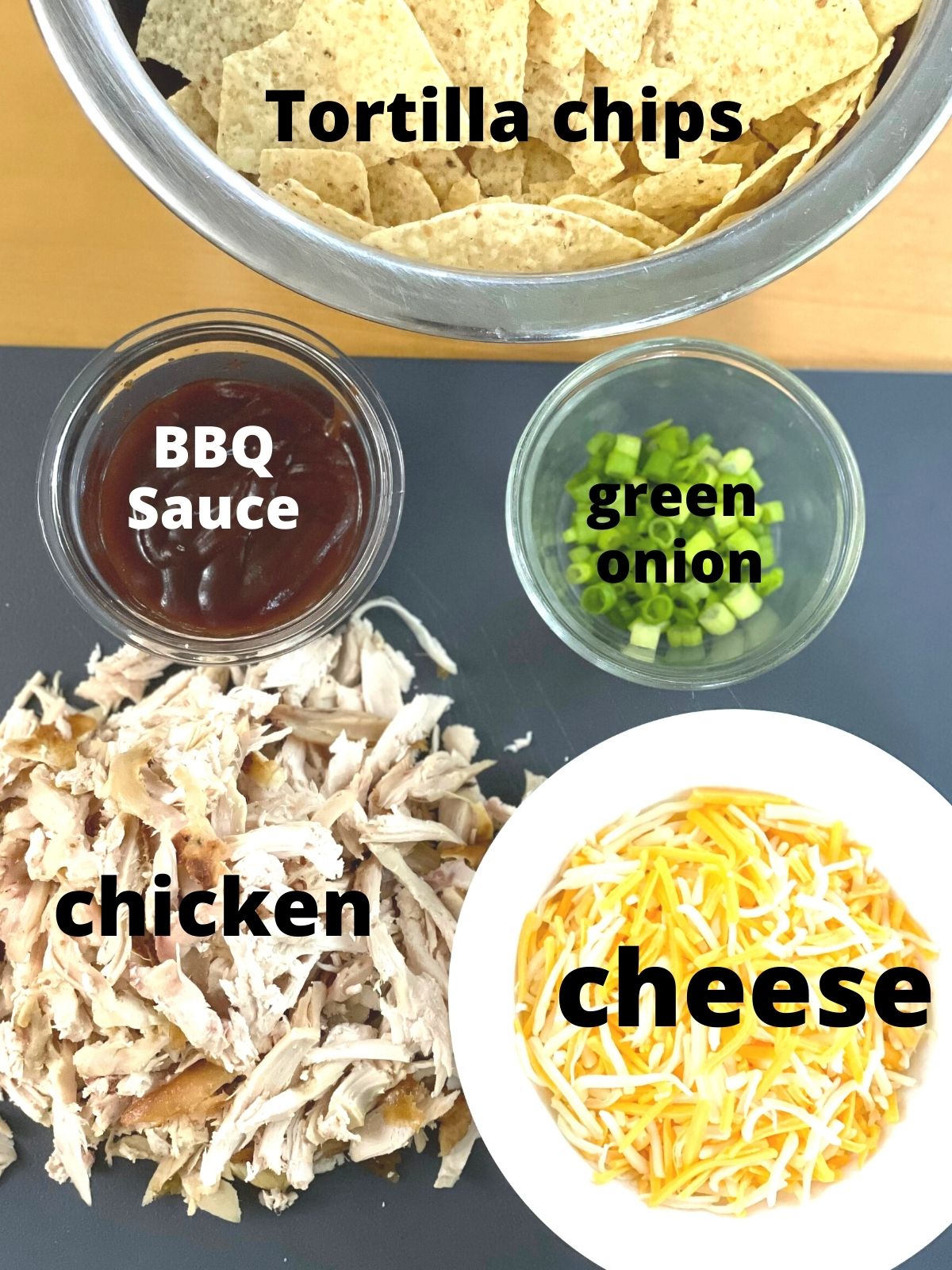 a bowl of tortilla chips surrounded by smaller bowls of nacho toppings as well as shredded chicken on a cutting board.
