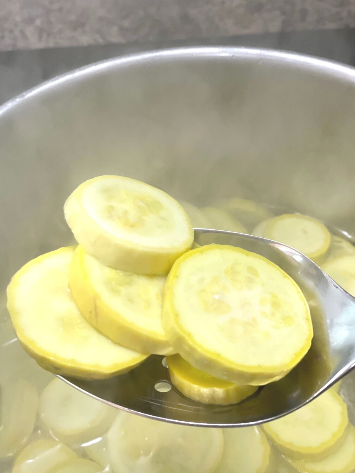 A slotted spoon is being use to remove yellow squash slices from the pot of boiling water.