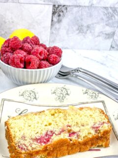 A slice of lemon loaf cake with raspberries on a plate. A bowl of fresh raspberries is in the background.