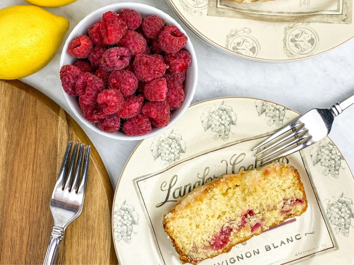 a slice of lemon loaf cake with raspberries in a bowl in the background.