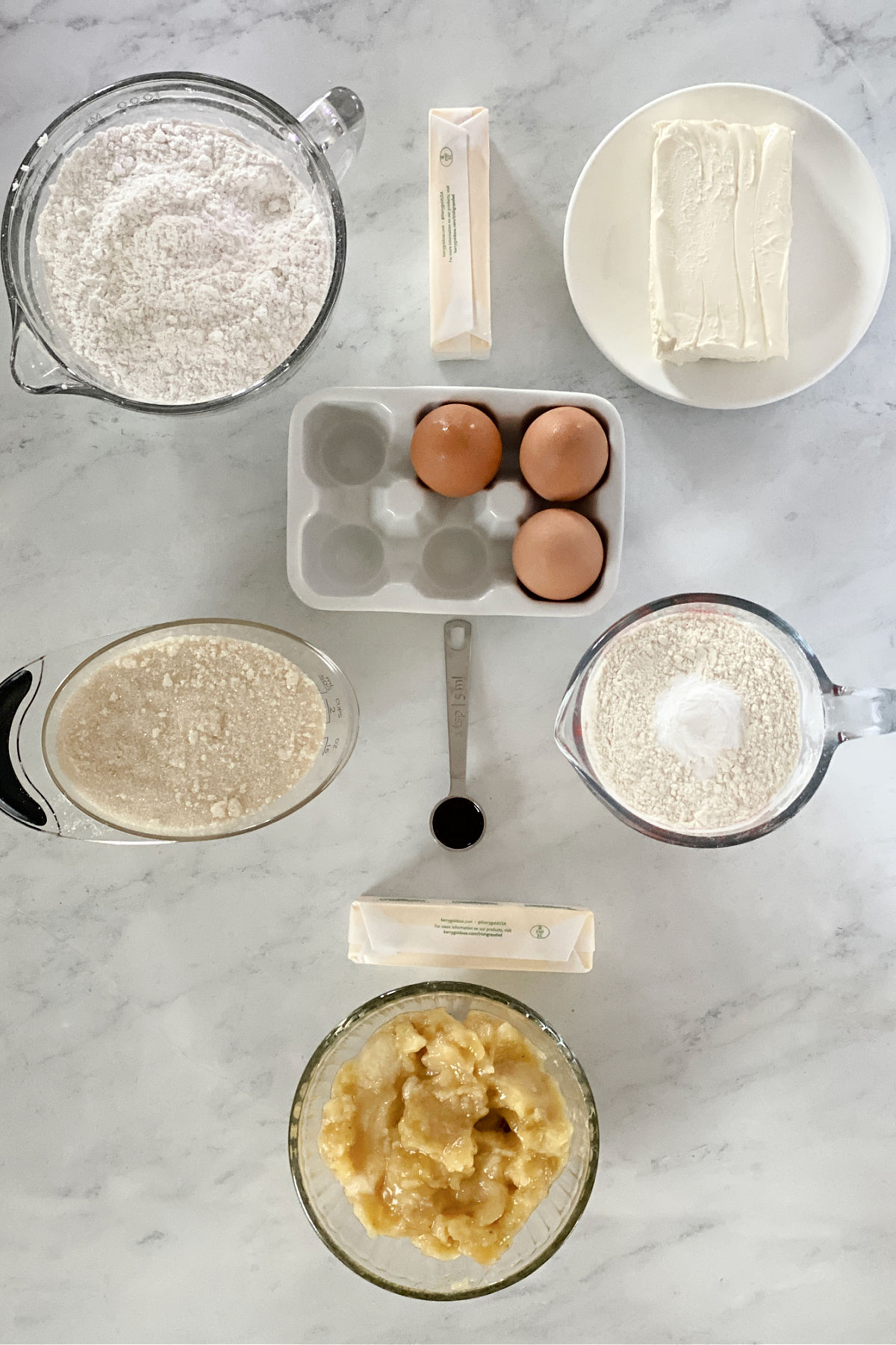 Flour, cream cheese, eggs, sugar, butter, vanilla, and mashed bananas on a counter top.