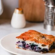 Freshly baked blueberry coffee cake square served on a white plate with utensils in the background.