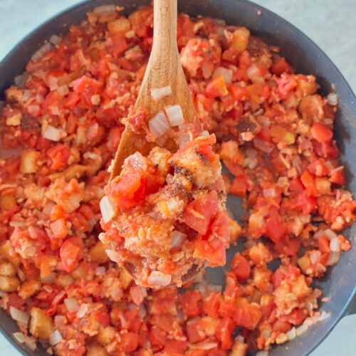 An iron skillet full of authentic Southern stewed tomatoes with a wooden spoon lifting a bite out of the pan.