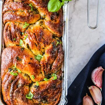 A loaf of homemade garlic bread still in the bread pan topped with fresh basil.