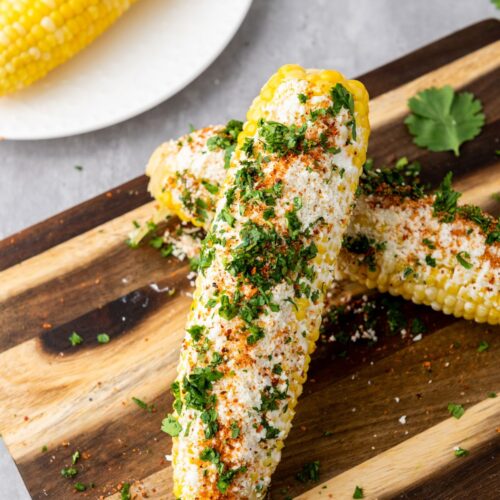 Two servings of Mexican elote arranged on a wooden cutting board and topped with cilantro.