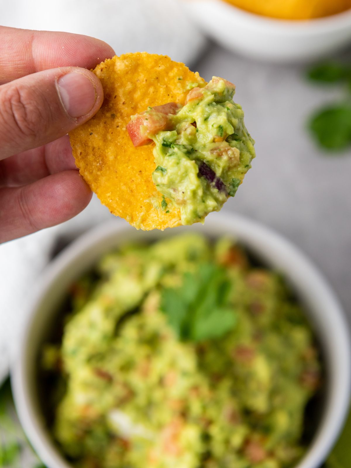 A chip full of guacamole held up to the camera with the bowl of guacamole in the background.