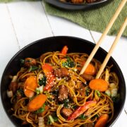 A black bowl full of pork lo mein topped with sesame seeds with chopsticks in the bowl.