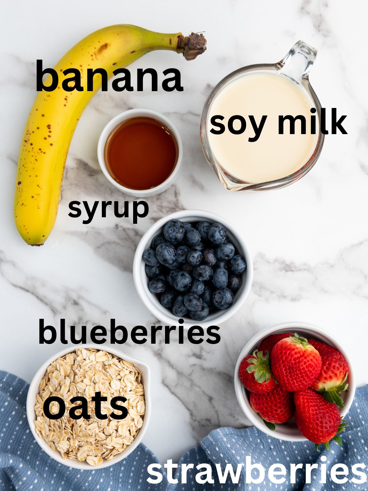 Vanilla overnight oats ingredients laid out on a kitchen counter: banana, soy milk, syrup, blueberries, oats, strawberries.